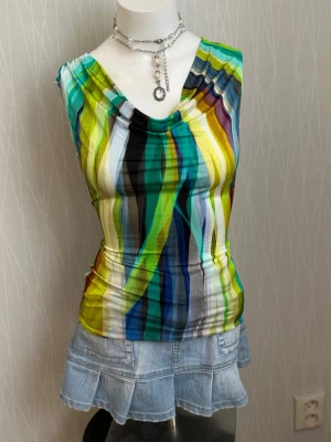 Flerfärgad randig ärmlös topp - Säljer en färgglad ärmlös topp med randigt mönster i grönt, blått, gult, svart och vitt. Toppen har en snygg draperad v-ringning och är figurnära i modellen. Perfekt för dig som vill sticka ut med en unik och trendig stil.