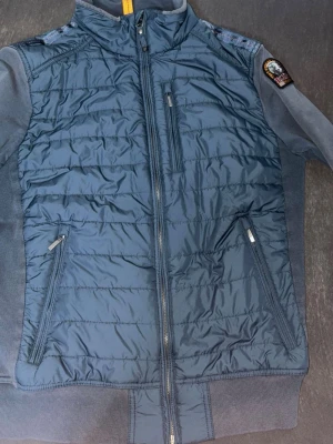 Mörkblå dunväst från Parajumpers - Snygg mörkblå dunväst från Parajumpers med quiltad framsida, hög krage och dragkedja. Västen har två snedställda fickor med dragkedja och en bröstficka. Märkets patch syns på axeln och detaljer i gult vid halsen. Perfekt för lager på lager.