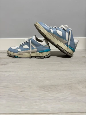 Blå och vita sneakers från Axel Arigato - Snygga sneakers från Axel Arigato i vitt och ljusblått läder med detaljer i turkos. Klassisk låg modell med perforerad tå, vita skosnören och robust sula. Märkeslogga på sidan och plös. Perfekta för en clean och trendig streetstil.