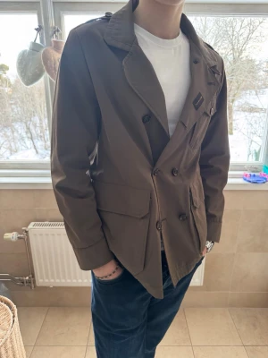 Brun rock från MooRER - Snygg brun rock från MooRER med dubbelknäppning och klassisk krage. Jackan har stora fickor framtill och en diskret logotyp på bröstet. Perfekt för dig som vill ha en stilren och tidlös look till vardags.