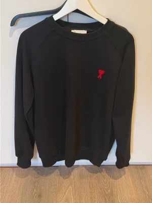 Svart sweatshirt från Ami Paris - Svart sweatshirt från Ami Paris med klassisk rund halsringning och lång ärm. På bröstet finns Ami Paris ikoniska röda hjärta med A-broderi. Tröjan har ribbade muddar vid ärmslut och nederkant. Perfekt för en clean och stilren look.
