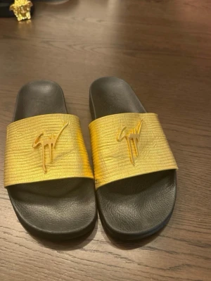 Guldiga slides från Giuseppe Zanotti - Säljer ett par snygga slides från Giuseppe Zanotti med bred guldfärgad rem och ikonisk logga i guld. Sulan är svart och har en bekväm formad fotbädd. Perfekta för en trendig och avslappnad stil.