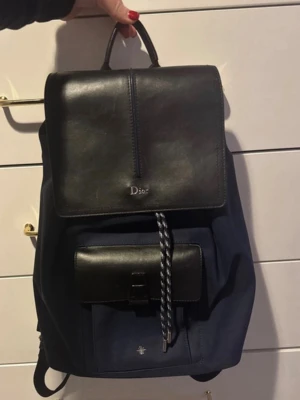 Mörkblå och svart ryggsäck från Dior - Snygg ryggsäck från Dior i mörkblått tyg med svarta läderdetaljer. Väskan har ett lock i läder med Dior-logga, en praktisk ficka framtill och justerbara axelband. Perfekt för dig som vill ha en stilren och exklusiv look.