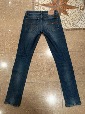 Nudie Jeans w30L32  - Säljer ett par klassiska blå jeans från Nudie Jeans med smal passform och snygga slitningar. Jeansen har fem fickor, orange sömmar och är tillverkade i denim. Perfekta för en avslappnad och trendig look.