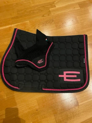 Svart schabrak & huva med rosa kant - Säljer ett svart schabrak och matchande huva från Equestrian Stockholm. Båda har rosa kant och logga i rosa. Schabraket har bikakemönster och är vadderat, perfekt för ridning. Materialet är textil och mesh. Har använt det få tal gånger. Super fint skick! Stolek PONNY!