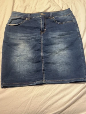 Blå jeanskjol med fickor - Snygg blå jeanskjol i klassisk modell med fem fickor och knapp framtill. Kjolen har en kort längd och är tillverkad i denim med ljusa slitningar för en trendig look. Perfekt att matcha med en t-shirt eller hoodie.