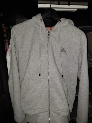Grå zip hoodie från Burberry - Snygg grå hoodie från Burberry med dragkedja och klassiskt rutigt mönster i huvan. Hoodien har dragsko i huvan, fickor framtill och en liten Burberry-logga broderad på bröstet. Storlek S använd några gånger men inga spår av användning.