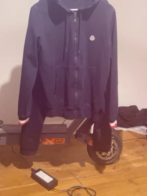 Mörkblå hoodie från Moncler - Snygg mörkblå hoodie från Moncler med dragkedja framtill och klassisk Moncler-logga på bröstet. Huva med dragsko och fickor på magen. Röd och vit rand vid ärmslut och halsringning ger en sportig touch. Perfekt för dig som gillar streetwear och exklusiva märken.