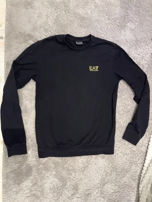 Svart EA7 Emporio Armani sweatshirt - Svart sweatshirt från EA7 Emporio Armani med rund halsringning och gul logotyp på bröstet. Tröjan har långa ärmar och ribbade muddar vid ärmslut och nederkant. Perfekt för en stilren och sportig look.