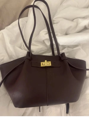 axel väska från gina tricot  - Snygg handväska i slätt läder med två långa axelband och dekorativt guldfärgat spänne framtill. Väskan har en modern, vingeformad siluett och stilrena linjer. Perfekt för dig som gillar minimalistisk design och lyxiga detaljer.