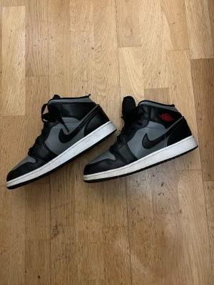 Air Jordan 1 Mid Shadow svart/grå - Snygga Air Jordan 1 Mid sneakers i svart och grått läder med klassisk svart swoosh och vita sulor. Röd logga på sidan och tungan ger extra detaljer. Säljer för dem är för små. Köpta på kicks n stuff i Göteborg. Utan låda.   NYPRIS 3000