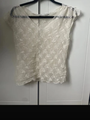 Vit spets-topp med v-ringning - Säljer en söt vit topp i genomskinlig spets med v-ringning och korta ärmar. Toppen har ett romantiskt blommönster och är perfekt att styla lager på lager. Materialet är tunt och lätt, vilket ger en luftig känsla.