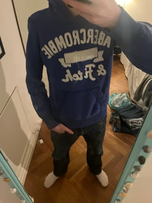 Blå hoodie från Abercrombie & Fitch - Snygg blå hoodie från Abercrombie & Fitch med stor vit logga framtill. Klassisk känguruficka och huva med dragsko. Perfekt för en avslappnad och cool stil. Mjuk bomullsblandning som är skön att ha på sig. Det står M på tröjan men skulle defenitivt säga att den är storlek xs-s 