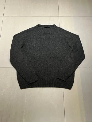 Mörkgrå stickad tröja med armbågslappar - Mörkgrå stickad tröja i klassisk modell med rund halsringning och ribbade muddar. Sitter som M, även om det står L på tröjan!!