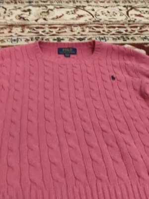 Rosa kabelstickad tröja Polo Ralph Lauren - Rosa kabelstickad tröja från Polo Ralph Lauren med rund halsringning och klassisk blå logga broderad på bröstet. Tröjan har ribbstickade muddar och är perfekt för dig som gillar en preppy stil.