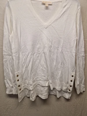 Vit tröja med knappar Michael Kors  - Vit stickad tröja med V-ringning och långa ärmar. Tröjan har dekorativa guldfärgade knappar på båda sidor vid nederkanten och en snygg lager-på-lager-effekt. Perfekt för dig som gillar en clean och enkel stil.
