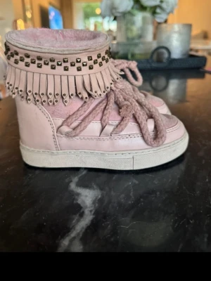 Rosa boots med fransar från Inuikii storlek 28 - Snygga rosa boots från Inuikii med fransar och nitar upptill. Skorna har snörning framtill och är fodrade med mjukt material. Perfekta för dig som vill ha något unikt och trendigt på fötterna under kallare dagar.