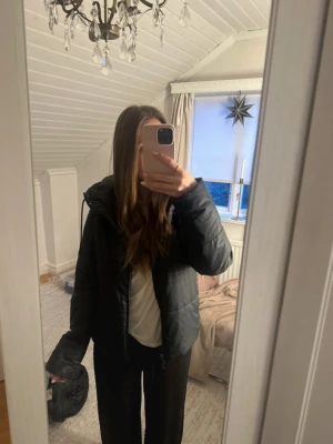 Svart pufferjacka från Nike - Säljer en svart pufferjacka från Nike med huva och dragkedja. Jackan har en clean look med små Nike-loggor på insidan av huvan. Perfekt för kalla dagar och riktigt skön att bära. Passformen är normal och jackan är lätt att matcha med olika outfits.