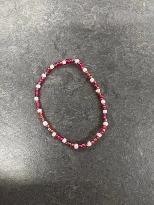 Färgglatt pärlarmband i rött och vitt Storlek M - Ett handgjort armband med små pärlor i rött och vitt. Pärlorna är trädda på en elastisk tråd vilket gör det enkelt att ta på och av. Perfekt accessoar för att addera lite färg till din stil.