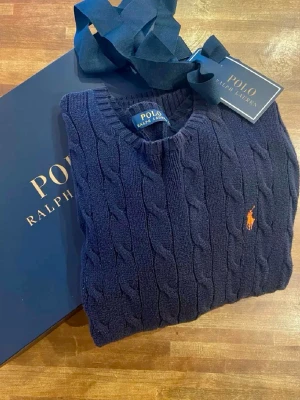 Mörkblå kabelstickad tröja Polo Ralph Lauren - Klassisk kabelstickad mörkblå tröja från Polo Ralph Lauren med rund halsringning och klassisk röd broderad logga på bröstet. 