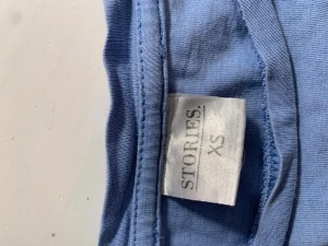 Ljusblå t-shirt från Stories XS - En enkel och stilren ljusblå t-shirt från Stories i storlek XS. T-shirten har klassisk passform med korta ärmar och rund halsringning. Perfekt basplagg att matcha med jeans eller kjol.