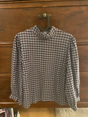 Rutig blus med hög krage - Snygg blus med brunt och vitt rutmönster och hög krage. Blusen har puffiga långärmar med resår vid ärmslut och en avslappnad passform. Perfekt för dig som gillar retrovibbar och vill sticka ut lite extra.