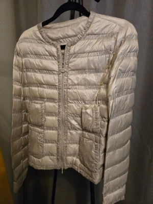 Moncler "Lissy giubbotto" Beige dunjacka med dragkedja - Stilren beige dunjacka från Moncler "Lissy giubbotto" med quiltat mönster och rund halsringning. Jackan har fyra framfickor och stängs med dragkedja framtill. Materialet är glansigt och lätt, perfekt för både vår och  vinterdagar. Passar dig som gillar en enkel men modern look. Som ny!