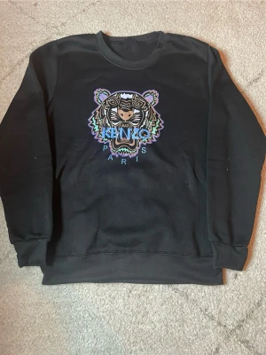  Kenzo sweatshirt - Cool svart sweatshirt från Kenzo med ikoniskt tigerhuvud broderat framtill i färgerna lila, blått, grönt och vitt. Rund halsringning och ribbade muddar vid ärmslut och nederkant. Perfekt statement-plagg för dig som gillar streetwear.