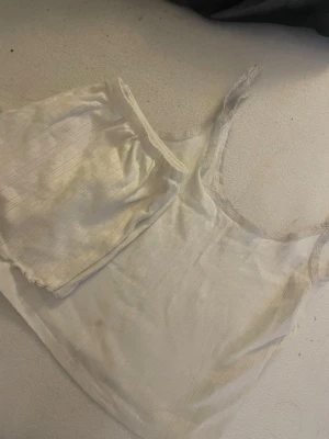 Vitt ribbat linne med spetsdetaljer - Säljer ett vitt loungewear-linne med tunna axelband och spetsdetaljer längs kanterna. Tyget är ribbat med diskreta ränder och har en mjuk, bekväm känsla. Perfekt för att chilla hemma eller som ett lager under andra plagg.