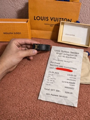 Louis Vuitton svart läderarmband - Stilrent svart armband från Louis Vuitton i präglat läder med guldfärgad LV-logga framtill. Snygg och tidlös accessoar som lyfter din outfit. Kommer med originalförpackning och dustbag.