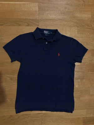 Marinblå pikétröja från Ralph Lauren - Klassisk marinblå pikétröja från Ralph Lauren med korta ärmar och röd broderad logga på bröstet. Tröjan har krage och knäppning framtill. Perfekt för en stilren och avslappnad look.