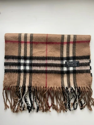 Beige rutig halsduk från Burberrys - Klassisk beige halsduk från Burberrys i 100% kashmir. Den har det ikoniska rutmönstret i beige, svart, vitt och rött samt fransar i ändarna. Märkt 'Made in England' och känns superlyxig och mjuk. Perfekt accessoar för att lyfta din outfit.