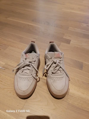Ljusrosa Adidas sneakers i mocka - Snygga rosa sneakers från Adidas i mjuk mocka med klassisk låg profil och gummisula. Skorna har diskret Adidas-logga på sidan och tungan samt perforerade detaljer för extra stil. Perfekta för dig som gillar en clean och stilren look.