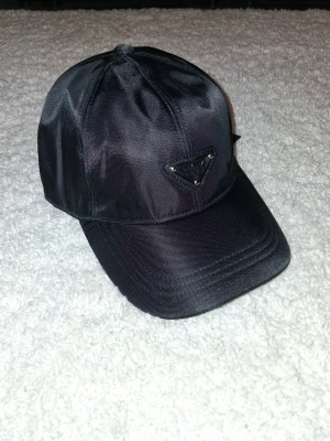 Svart keps från Prada i nylon - Snygg svart keps från Prada i glansigt nylonmaterial med klassisk böjd skärm. Framtill sitter Pradas ikoniska trekantslogga i metall. Perfekt accessoar för en trendig och stilren look.