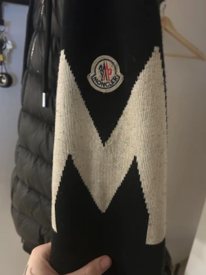 Svart hoodie från Moncler med mönster - Säljer en svart hoodie från Moncler med stor vit zigzag-design på ärmarna och Moncler-logga på ena ärmen. Hoodien har en klassisk huva och är gjord i mjukt material som känns riktigt skönt. Perfekt för dig som vill ha något stilrent men ändå sticker ut. Äkta qr och scann finns 