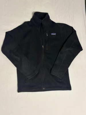 Svart patagonia fleece - Mysig svart fleecejacka från Patagonia med hel dragkedja, hög krage och bröstficka med dragkedja. Perfekt för lager-på-lager och har en enkel, stilren design. Materialet är mjukt och värmande, och jackan har två sidofickor. Storlek XS