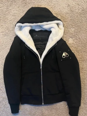 Svart dunjacka från Moose Knuckles - Snygg svart dunjacka från Moose Knuckles med vit fuskpäls i huvan och quiltad insida. Jackan har dragkedja framtill, två stora fickor och en ikonisk logga på ärmen. Perfekt för kalla vinterdagar och har en riktigt clean look. 