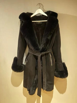Svart fuskpäls kappa  - Snygg svart pälsjacka från Gloria Fashion med stor fluffig huva, markerade ärmslut och kant i fuskpäls. Den har midjebälte och dragkedjor i silver på sidorna. Passar strl S-L