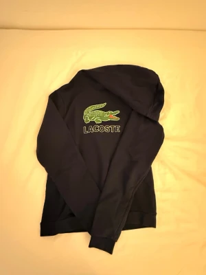 Svart hoodie från Lacoste med logga - Snygg svart hoodie från Lacoste med den klassiska gröna krokodilloggan och texten 'LACOSTE' broderad på bröstet. Hoodien har huva och ribbade muddar. Perfekt för dig som gillar streetwear och vill ha en stilren look.
