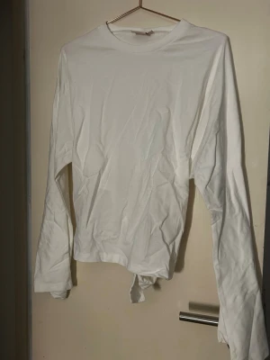 Vit långärmad topp med knyt i ryggen - Snygg vit långärmad topp med rund hals och unik knyt-detalj i ryggen. Toppen är croppad och har en clean look som passar till mycket. Tillverkad i mjukt bomullstyg för en bekväm känsla. Ny med prislapp