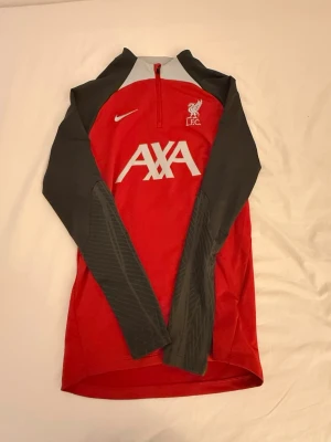 Liverpool Nike röd träningsjacka - Röd träningsjacka från Nike med Liverpool FC-logga och AXA-tryck på bröstet. Jackan har gråa ärmar, halv dragkedja och sportig design. Perfekt för fotbollsträning eller som supporterplagg. Tillverkad i funktionsmaterial för bra komfort.