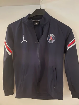 PSG x Jordan mörkblå träningsset - Snyggt mörkblått träningsset från PSG x Jordan med jacka och byxor. Setet har vita och röda detaljer på ärmar och ben, PSG-logga och Jumpman-logga på både jacka och byxor. Perfekt för fotbollsträning eller chill. Materialet är mjukt och stretchigt. Drag kedjan på koftan saknar en liten metall bit så den är svår att stänga och hålla ihop men den kan ändå funka.