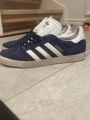 Adidas Gazelle blå och vita sneakers - Klassiska Adidas Gazelle sneakers i mörkblå mocka med vita läderdetaljier. Jag säljer dem för att jag inte vill ha dem längre och säljer dem för ett riktigt bra pris. 