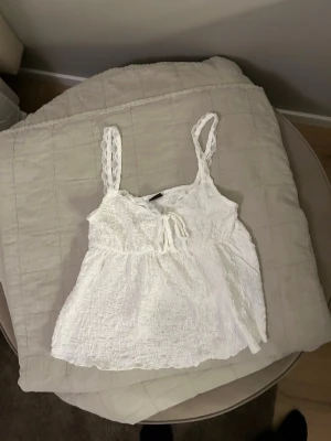 Vit spetslinnetopp från Gina Tricot - Söt vit linnetopp från Gina Tricot med spetsdetaljer och knyt framtill. Toppen har tunna axelband i spets och ett lätt skrynklat tyg som ger en avslappnad känsla. Perfekt för varma dagar och somriga outfits. Använd 1 gång