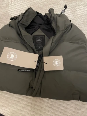 Mörkgrön pufferjacka från Canada Goose - Säljer en mörkgrön pufferjacka från Canada Goose med huva och svarta detaljer. Jackan har dragkedja framtill, två sidofickor och patch med logga på ärmen. Perfekt för kalla dagar och riktigt snygg till streetwear-stilen.