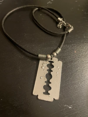 Silvrigt halsband med rakbladshänge - Coolt halsband med ett hänge i form av ett silvrigt rakblad. Kedjan är tunn och metallisk, och hänget har en klassisk rakbladsdesign som ger en edgy känsla. Perfekt accessoar för dig som gillar alternativ stil.