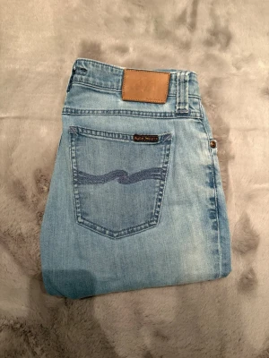 Ljusblå jeans från Nudie Jeans - Säljer ett par ljusblå jeans från Nudie Jeans med klassisk femficksmodell och raka ben. Jeansen har en snygg tvättad look, läderpatch bak i midjan och broderad bakficka. Perfekta för dig som gillar avslappnad stil. Storlek:30-32.   Innerbensläng:74cm, ytterbenslängd:97cm, midjevidd:40cm, benvidd:14cm.
