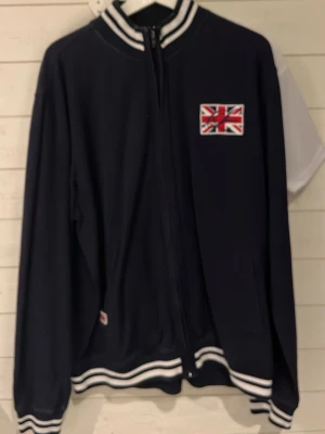 Mörkblå ziptröja med brittisk flagga - Snygg mörkblå ziptröja med Union Jack-flagga på bröstet och vita ränder på krage, ärmslut och midja. Tröjan har sportig collegekänsla och är perfekt för dig som gillar streetwear. Långärmad och med dragkedja framtill.