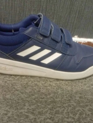 Blå Adidas sportskor med kardborre - Blå sportskor från Adidas med tre vita ränder på sidan och praktiska kardborreband. Skorna har en vit sula och ovandel i syntetmaterial och textil. Perfekta för träning eller en aktiv vardag.