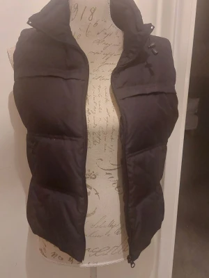 Svart dunväst med hög krage - Snygg svart dunväst med dragkedja och hög krage. Västen har en puffig design och justerbar dragsko vid halsen. Perfekt för lager-på-lager och passar till många olika outfits. Materialet är mjukt och håller värmen bra. Lite liten I storleken.